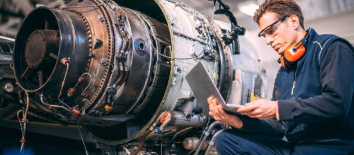 Aircraft engineer in a hangar holding laptop computer while repairing and maintaining a jet engine.